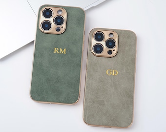 Op de afbeelding: Twee olijfgroene telefoonhoesjes met goudkleurige randen en camera-omlijstingen. Elke hoes heeft een gouden monogram, één met "RM" en de andere met "GD". De hoesjes liggen op een wit oppervlak in de buurt van een laptop.