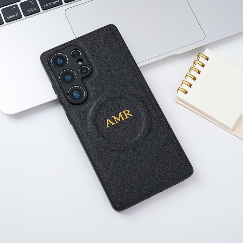 May include: Black leather phone case with a textured finish. The case features a circular design with the gold initials "AMR". The phone case is lying on a white surface next to a laptop and a small notepad.