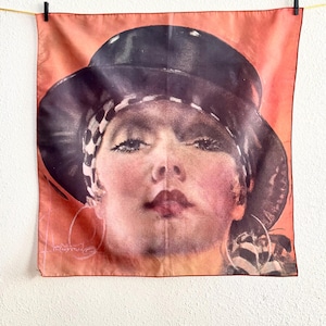 May include: A vintage silk scarf featuring a portrait of a woman with a black hat and hoop earrings. The woman's face is painted with pink blush and red lipstick. The scarf is peach-colored and is hanging from a clothesline.