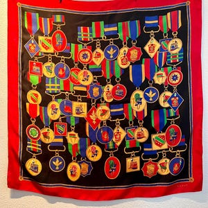 Puede incluir: Un pañuelo cuadrado negro con un borde rojo y un patrón repetido de medallas y cintas coloridas. Las medallas doradas tienen varios diseños, y las cintas son rojas, verdes y azules. El pañuelo está colgado de un tendedero.