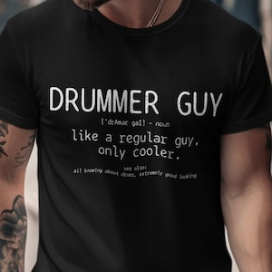 Può includere: T-shirt nera con la scritta bianca "DRUMMER GUY" e una definizione. La definizione recita "come un ragazzo normale, solo più cool". La maglietta è a maniche corte ed è indossata da una persona.