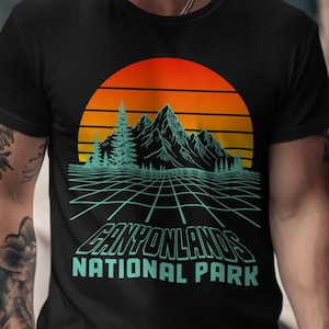 May include: Black t-shirt featuring a retro design of a sunset, mountains, and grid lines. The text "Canyonlands National Park" is displayed in a teal font, evoking a vintage aesthetic. A classic tee for outdoor enthusiasts.