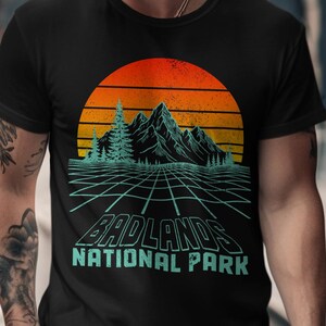May include: Black t-shirt featuring a retro design with a sunset, mountains, and the text "BADLANDS NATIONAL PARK" in teal. The graphic has a grid-like pattern. A classic crew neck tee.