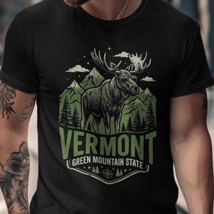 May include: Black t-shirt featuring a graphic of a moose with large antlers, set against a backdrop of green mountains and trees. The word "VERMONT" is prominently displayed, with "GREEN MOUNTAIN STATE" below.