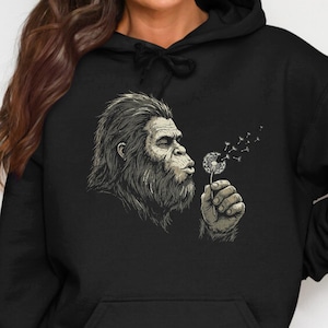 May include: A black hooded sweatshirt featuring a graphic illustration of a hairy, ape-like creature blowing on a dandelion. The creature is depicted in shades of grey and white, with the dandelion seeds drifting away into the air.