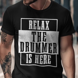 Può includere: T-shirt nero con la scritta "RELAX THE DRUMMER IS HERE" in bianco, con un carattere invecchiato. Il testo è centrato all'interno di una cornice rettangolare bianca. La maglietta è indossata da una persona.