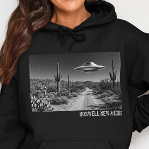 May include: A black hooded sweatshirt featuring a rectangular graphic print of a desert landscape with saguaro cacti, a flying saucer, and a small figure. The text ROSWELL NEW MEXICO is printed in white capital letters below the graphic.