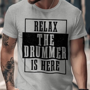Può includere: T-shirt grigio chiaro con la scritta "RELAX THE DRUMMER IS HERE" in nero e in grassetto. Il testo è racchiuso in una cornice rettangolare nera. La maglietta è a maniche corte.