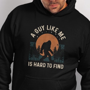 May include: Black hooded sweatshirt with a Bigfoot graphic. The design features a silhouette of Bigfoot in front of an orange sun, with the text "A Guy Like Me Is Hard To Find". The sweatshirt has a front pocket and a drawstring hood.