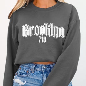 May include: A dark gray sweatshirt with the word "Brooklyn" in a white, bold, stylized font across the chest. Below the word "Brooklyn" is the number "718" in white. The sweatshirt is paired with blue jeans.