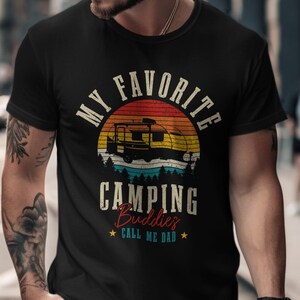 May include: Black t-shirt with a camping-themed graphic. The design features a camper silhouette against a sunset backdrop, with the text "MY FAVORITE CAMPING BUDDIES CALL ME DAD". The design is in a vintage style.