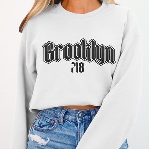 May include: White sweatshirt with the word "Brooklyn" in a black gothic font, with "718" below it. The sweatshirt is worn with blue jeans.