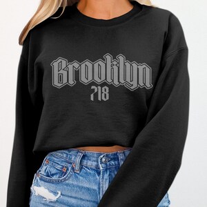 May include: Black crew neck sweatshirt with the word "Brooklyn" in a gray, stylized font, and the number "718" below it. The sweatshirt is worn with blue jeans.