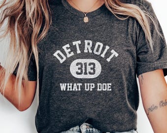 May include: Dark gray t-shirt with the text "DETROIT 313 WHAT UP DOE" in white. The shirt has a crew neck and short sleeves. The person is wearing light blue jeans.