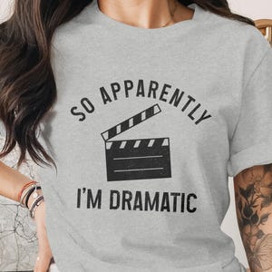 May include: A pale grey t-shirt featuring the words "SO APPARENTLY I'M DRAMATIC" in black, above a black film clapperboard graphic. The shirt has short sleeves and a crew neck.