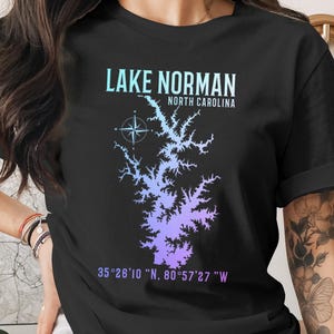 May include: A black short-sleeve t-shirt featuring a light blue to purple gradient map of Lake Norman, North Carolina. The graphic includes a compass rose, the text "LAKE NORMAN NORTH CAROLINA," and the coordinates "35°26'10 "N, 80°57'27 "W."