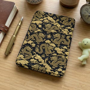 May include: A black and gold dragon pattern notebook lies on a wooden surface, next to a succulent plant in a patterned pot, a lit candle, a small alarm clock, and a small figurine. The notebook's design features gold dragons and clouds.