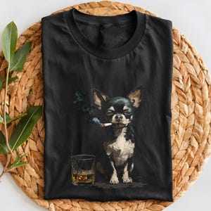May include: Black t-shirt featuring a Chihuahua dog with a cigarette in its mouth and a glass of whiskey. The dog is black and white with large ears. The whiskey glass is clear with amber liquid. The shirt is displayed on a woven, circular mat.