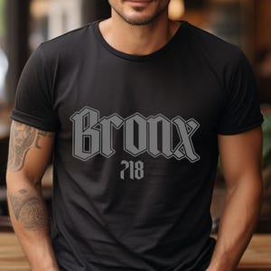 May include: Black t-shirt with the word "Bronx" in a gray, stylized font, and the number "718" below it. The shirt is a crew neck style.