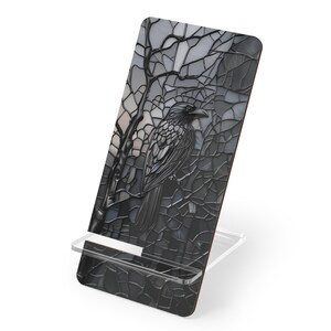 May include: A black phone stand featuring a raven design. The raven is perched on a tree branch, set against a stained-glass style background. The stand is supported by a clear acrylic base.