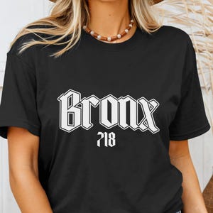 May include: Black t-shirt with the word "Bronx" in a bold, white, gothic font. Below the word "Bronx" is the number "718" in white. The t-shirt is a classic crew neck style.