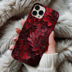 May include: A smartphone case featuring a vibrant floral design in shades of red. The case showcases a detailed pattern of stylized flowers with a dark red background. The phone case is held in a hand, with a white sweater sleeve visible.