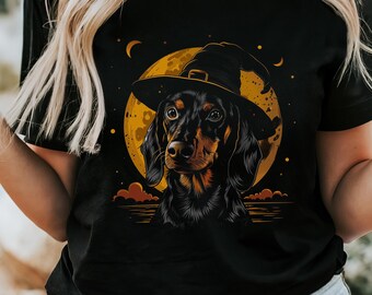 May include: A black short-sleeve t-shirt featuring a graphic print of a black and tan dachshund wearing a pointed witch hat. The dog is positioned in front of a large, circular yellow moon with small orange clouds and stars in the background.