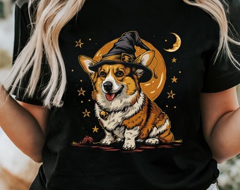 May include: A black t-shirt featuring a graphic illustration of a brown and white Corgi dog wearing a black witch hat. The dog is positioned in front of a large orange moon, surrounded by small yellow stars and a crescent moon in the night sky.