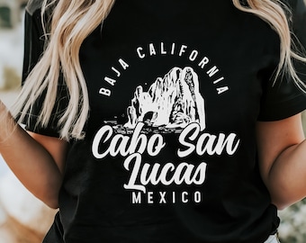 May include: Black t-shirt with a white graphic design featuring the text "BAJA CALIFORNIA Cabo San Lucas MEXICO". The design includes an illustration of rock formations. The shirt is worn with denim shorts.