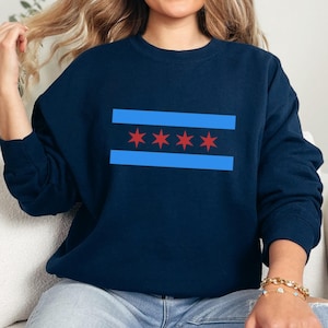 May include: Navy blue sweatshirt featuring the Chicago city flag design. The design includes two light blue horizontal stripes with four red stars in the center. The sweatshirt is a long-sleeved crew neck.