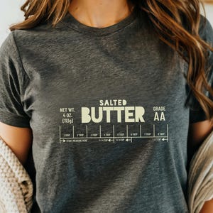 May include: A gray t-shirt with a graphic of a stick of butter with the text "SALTED BUTTER GRADE AA" and a measurement guide for tablespoons and cups. The text is in a light yellow color.