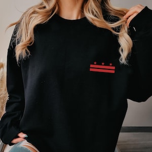 May include: Black sweatshirt with a red and white graphic design on the upper left chest. The design features two red horizontal lines and three red stars. The sweatshirt is long-sleeved.