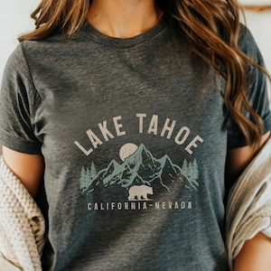 Puede incluir: Una camiseta gris oscuro con un gr&aacute;fico de estilo vintage de monta&ntilde;as, un oso y un sol. El texto "LAKE TAHOE CALIFORNIA-NEVADA" est&aacute; impreso en la camiseta.
