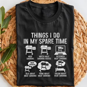 May include: Black t-shirt with white text and graphics about meat smoking. The text reads "THINGS I DO IN MY SPARE TIME" with illustrations of a smoker, laptop, and thought bubbles. The shirt is displayed on a woven mat.