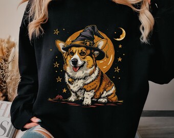 May include: A black long-sleeve sweatshirt featuring a graphic print of a brown and white Corgi dog wearing a black witch hat. The dog has a small bell collar and is positioned in front of a large orange moon, surrounded by yellow stars.