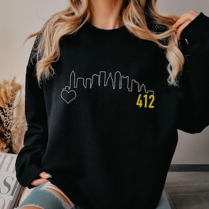 May include: Black sweatshirt featuring a white skyline graphic with a heart and the number "412" in yellow. The design is a minimalist outline of a city's buildings. The sweatshirt is a casual, comfortable style.