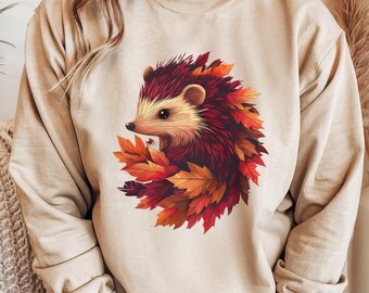 May include: Beige sweatshirt featuring a detailed illustration of a hedgehog surrounded by autumn leaves. The hedgehog has a reddish-brown back, a cream-colored face, and is nestled among red, orange, and yellow leaves. The sweatshirt has long sleeves.