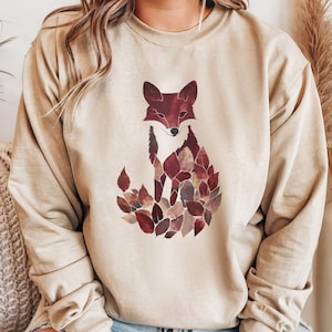 May include: Beige sweatshirt featuring a watercolor fox design. The fox is composed of maroon and brown leaves, with a matching maroon fox head. The long-sleeved top is a neutral color, suitable for fall.