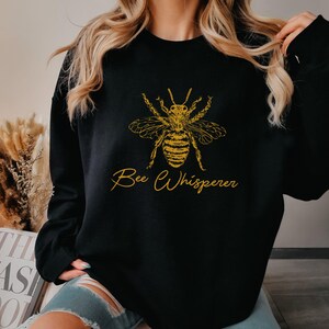May include: Black sweatshirt with a gold-colored bee graphic and the text "Bee Whisperer". The bee design is detailed, with visible wings and body segments. The sweatshirt is a casual style.