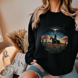 May include: Black sweatshirt with a graphic print of a woman riding a horse with a UFO in the background. The graphic is surrounded by a circle and has a green, brown, and orange color scheme.