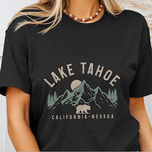Puede incluir: Camiseta negra con un dise&ntilde;o gr&aacute;fico vintage que presenta una cordillera, un oso y el texto "Lake Tahoe California-Nevada".