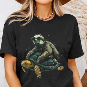 May include: Black t-shirt featuring a cartoon sloth wearing sunglasses riding on a turtle. The sloth is brown and green, and the turtle is yellow and green. The design is set against a dark background with white dots.