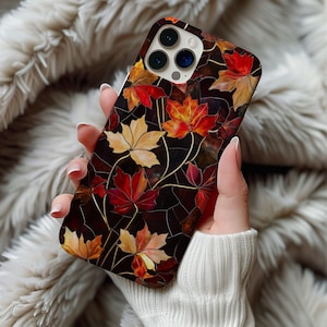 Puede incluir: Funda de teléfono con diseño de hojas de otoño. La funda tiene un fondo marrón oscuro con un patrón de hojas de arce rojas, naranjas y amarillas. Las hojas están dispuestas con vetas doradas, creando un efecto de mosaico. La funda del teléfono se sostiene en una mano.