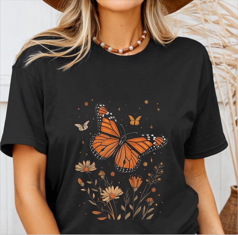 K&ouml;nnte beinhalten: Schwarzes T-Shirt mit einem Aufdruck eines orange-schwarzen Schmetterlings, umgeben von kleinen Blumen und Sternen.