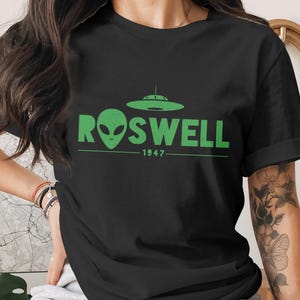 May include: A black short-sleeved t-shirt featuring a green graphic print on the chest. The design includes a flying saucer icon above the text ROSWELL, with an alien head replacing the letter O, and the year 1947 printed below the text.