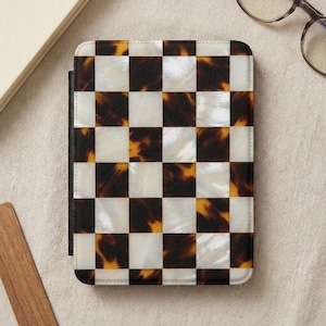 May include: A rectangular e-reader case featuring a checkered pattern of alternating white pearlescent squares and dark brown tortoiseshell squares. The case has a black spine on the left side and a textured finish across the entire front surface.