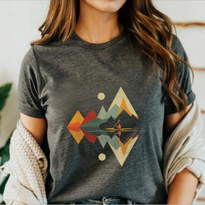 May include: A dark gray t-shirt with a geometric design of mountains and a reflection in water. The design features a canoe with two people paddling in the water. The mountains are in shades of orange, yellow, green, blue, and red.