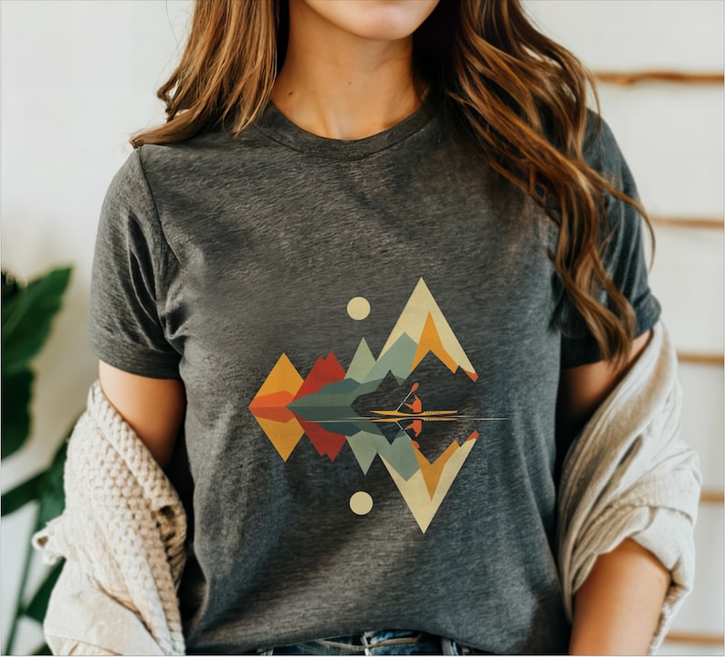 May include: A dark gray t-shirt with a geometric design of mountains and a reflection in water. The design features a canoe with two people paddling in the water. The mountains are in shades of orange, yellow, green, blue, and red.
