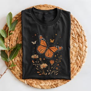 K&ouml;nnte beinhalten: Schwarzes T-Shirt mit einem Aufdruck eines orangefarbenen Schmetterlings, umgeben von gelben Blumen und goldenen Sternen.