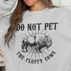 May include: White sweatshirt with a black and white graphic of a bison and the text "DO NOT PET THE FLUFFY COWS". The design features a detailed illustration of the animal with radiating lines.